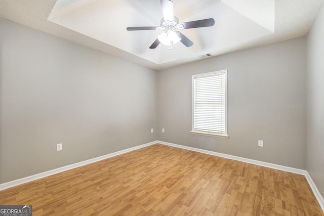 empty room with visible vents, a tray ceiling, light wood-style floors, baseboards, and ceiling fan