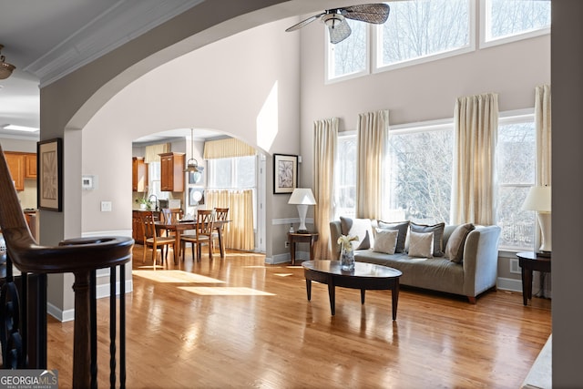 living area with light wood-style flooring, a ceiling fan, arched walkways, crown molding, and baseboards