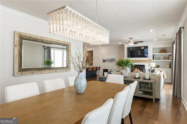 dining area featuring built in shelves, ornamental molding, a warm lit fireplace, wood finished floors, and ceiling fan