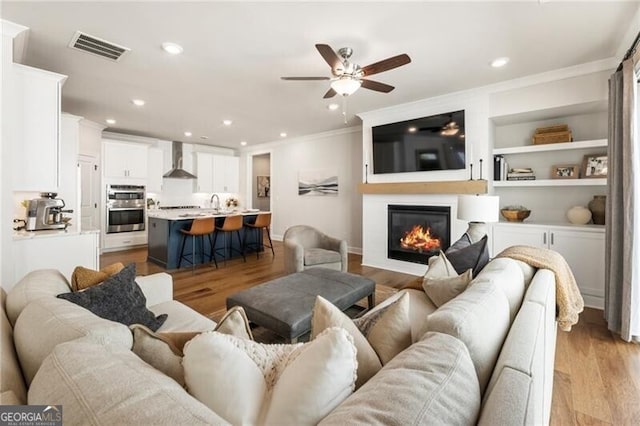 living room featuring light wood finished floors, visible vents, crown molding, and a glass covered fireplace