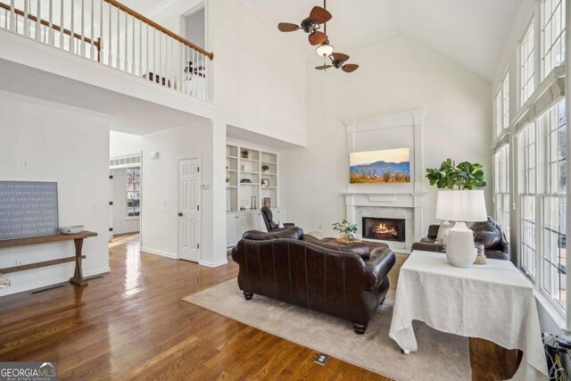 living area with built in shelves, wood finished floors, baseboards, a warm lit fireplace, and ceiling fan