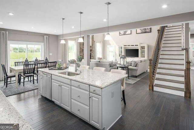 kitchen featuring dark wood finished floors, an island with sink, a sink, stainless steel dishwasher, and decorative light fixtures