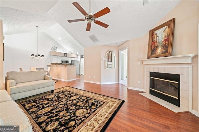 living area with a tiled fireplace, vaulted ceiling, ceiling fan with notable chandelier, wood finished floors, and a textured ceiling