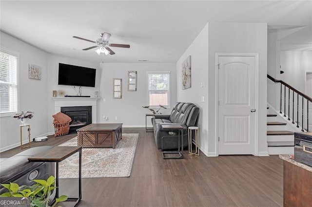 living room featuring a glass covered fireplace, stairway, wood finished floors, and a ceiling fan