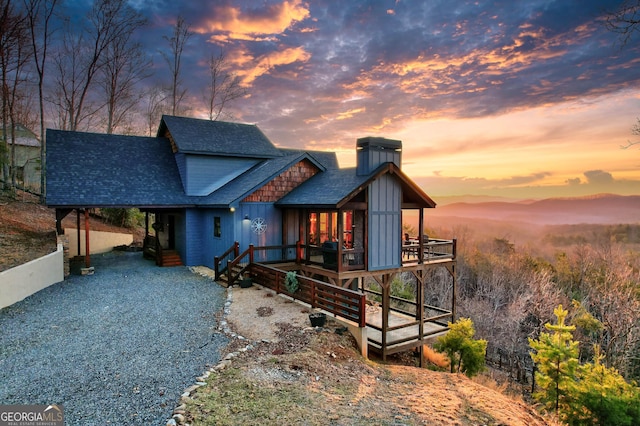 view of front of home featuring board and batten siding, a shingled roof, gravel driveway, a chimney, and a deck