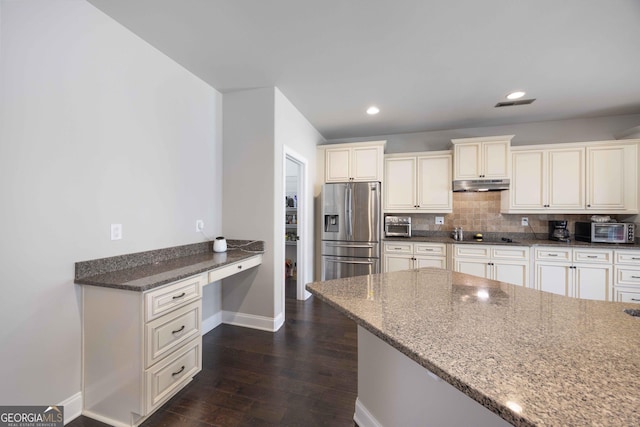 kitchen featuring under cabinet range hood, stone countertops, built in desk, stainless steel fridge with ice dispenser, and decorative backsplash