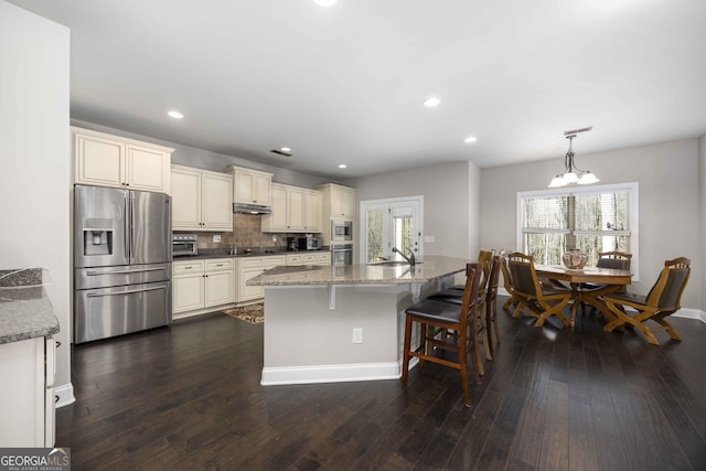 kitchen with dark wood finished floors, under cabinet range hood, appliances with stainless steel finishes, cream cabinets, and a sink