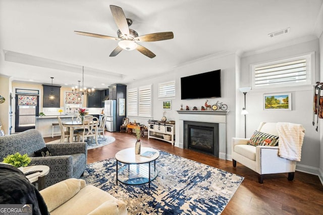 living area with visible vents, dark wood-type flooring, ornamental molding, ceiling fan with notable chandelier, and a glass covered fireplace