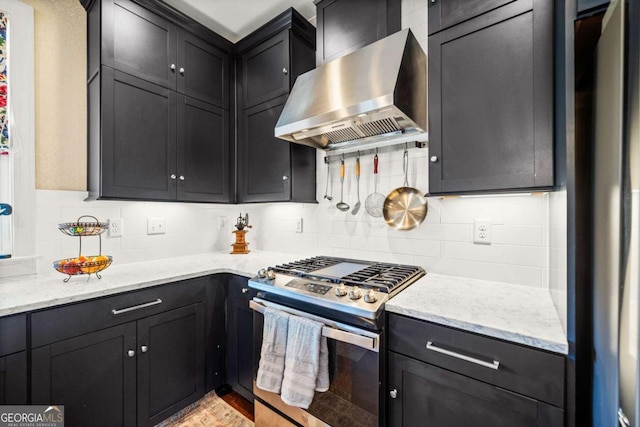 kitchen with dark cabinetry, light stone counters, appliances with stainless steel finishes, wall chimney range hood, and backsplash