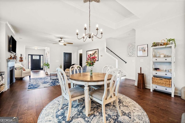 dining space featuring stairway, a fireplace, dark wood-style flooring, ornamental molding, and ceiling fan with notable chandelier