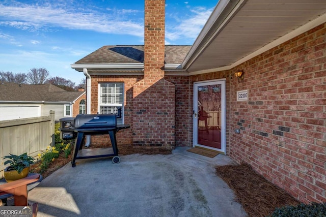 view of exterior entry featuring fence, a chimney, a shingled roof, a patio area, and brick siding