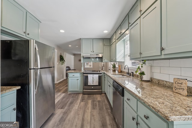 kitchen with light stone countertops, a sink, under cabinet range hood, appliances with stainless steel finishes, and backsplash