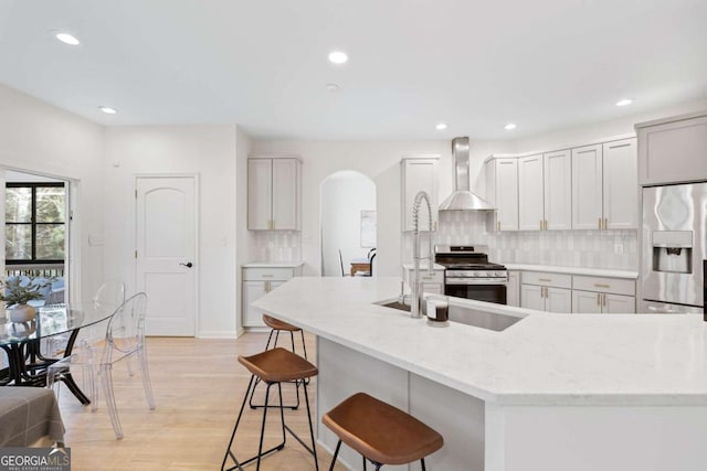 kitchen with tasteful backsplash, arched walkways, appliances with stainless steel finishes, a breakfast bar area, and wall chimney exhaust hood