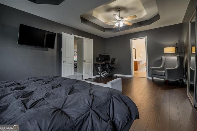 bedroom featuring baseboards, ensuite bathroom, wood finished floors, a raised ceiling, and a ceiling fan