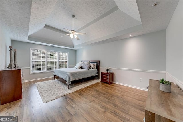 bedroom featuring a raised ceiling, a ceiling fan, a textured ceiling, wood finished floors, and baseboards