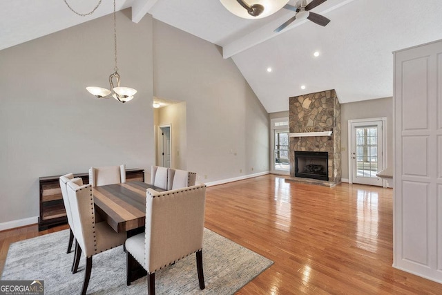 dining area featuring baseboards, a stone fireplace, light wood-style floors, beamed ceiling, and ceiling fan with notable chandelier