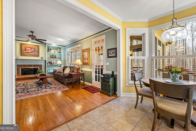 living room featuring a brick fireplace, built in shelves, ornamental molding, ceiling fan with notable chandelier, and wood-type flooring