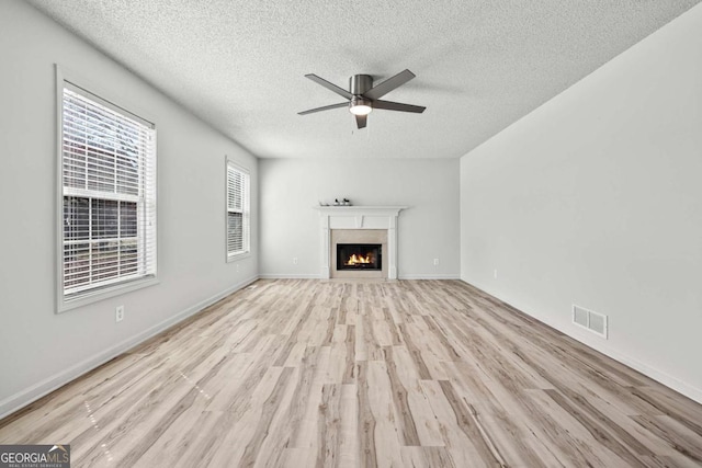 unfurnished living room featuring visible vents, a textured ceiling, a warm lit fireplace, light wood-style floors, and ceiling fan