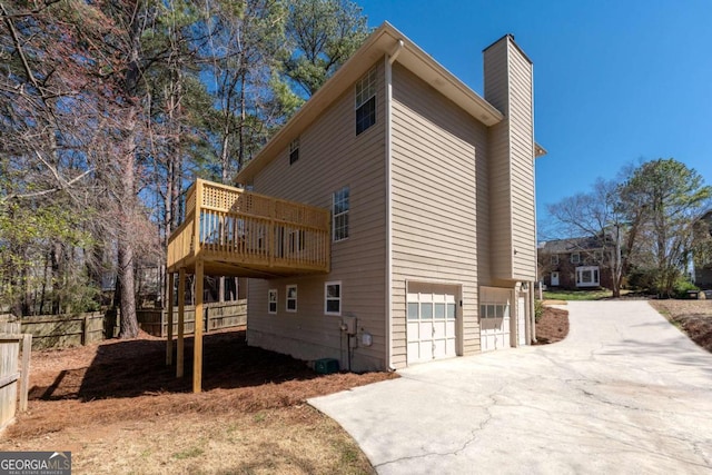 rear view of house featuring fence, concrete driveway, an attached garage, a wooden deck, and a chimney