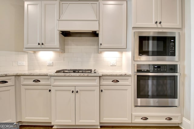 kitchen with light stone counters, stainless steel appliances, decorative backsplash, custom range hood, and white cabinetry