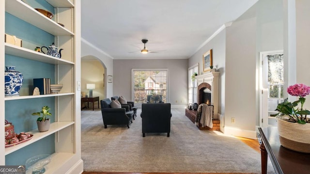 carpeted living area featuring ornamental molding, arched walkways, a lit fireplace, baseboards, and ceiling fan