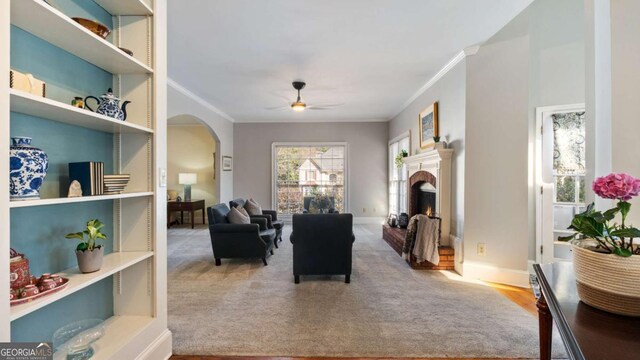 living area featuring a lit fireplace, crown molding, baseboards, arched walkways, and ceiling fan