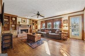 living room with built in shelves, crown molding, a warm lit fireplace, wood finished floors, and a ceiling fan