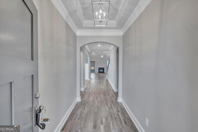 corridor featuring a raised ceiling, ornamental molding, wood finished floors, arched walkways, and baseboards