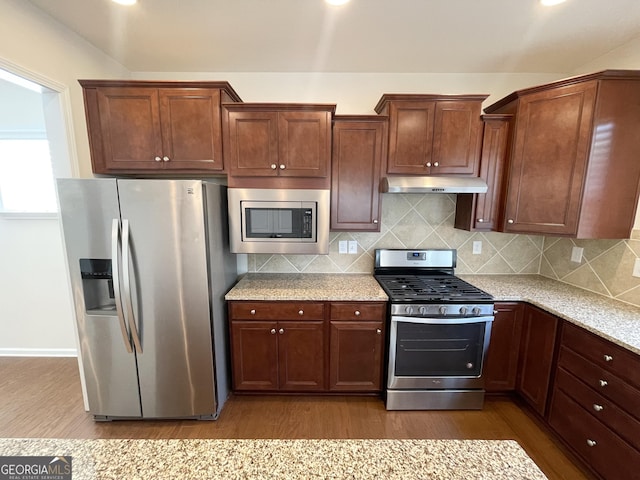 kitchen featuring under cabinet range hood, wood finished floors, appliances with stainless steel finishes, decorative backsplash, and light stone countertops