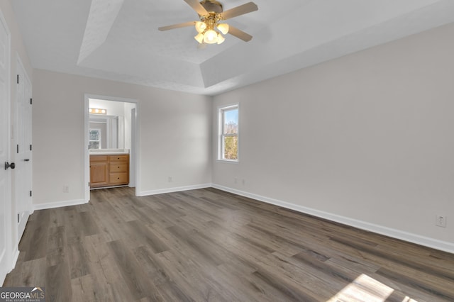 unfurnished bedroom featuring dark wood-type flooring, ceiling fan, baseboards, ensuite bath, and a raised ceiling