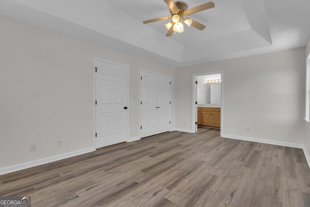 unfurnished bedroom featuring a raised ceiling, ensuite bath, wood finished floors, baseboards, and ceiling fan