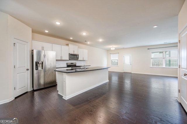 kitchen with dark wood-type flooring, a center island with sink, dark countertops, appliances with stainless steel finishes, and white cabinets