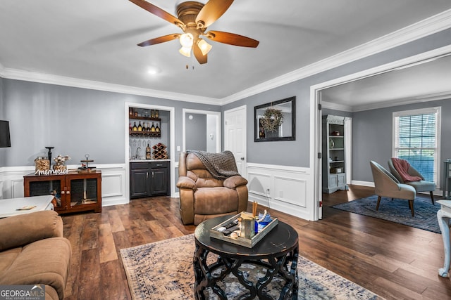 living area featuring dark wood-style floors, a ceiling fan, a wainscoted wall, crown molding, and a dry bar