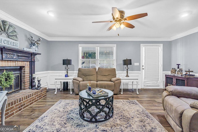 living area with a brick fireplace, ceiling fan, a wainscoted wall, ornamental molding, and dark wood-style floors