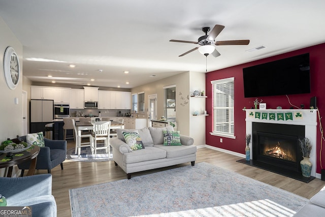 living area with a ceiling fan, visible vents, baseboards, a glass covered fireplace, and light wood-type flooring