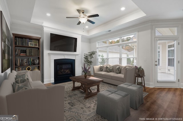 living area with wood finished floors, a fireplace with flush hearth, ceiling fan, crown molding, and a raised ceiling