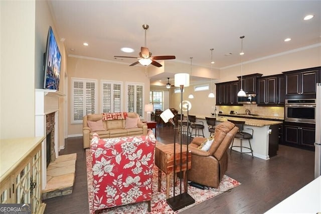 living area featuring a fireplace with raised hearth, ornamental molding, recessed lighting, a ceiling fan, and dark wood-style flooring