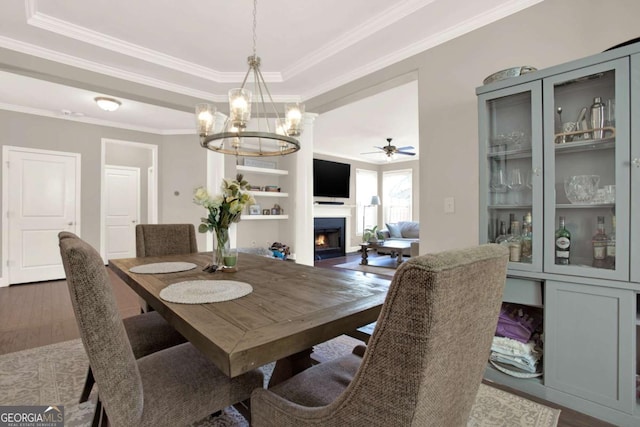 dining room with a tray ceiling, ceiling fan with notable chandelier, dark wood-type flooring, and a warm lit fireplace