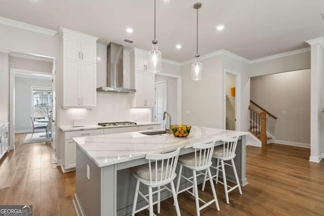 kitchen with stainless steel gas cooktop, an island with sink, white cabinetry, wall chimney exhaust hood, and a sink