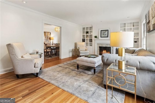 living room with built in shelves, ornamental molding, wood finished floors, a lit fireplace, and baseboards