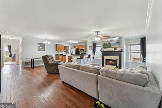 living area with visible vents, dark wood-style flooring, ornamental molding, a tile fireplace, and ceiling fan with notable chandelier