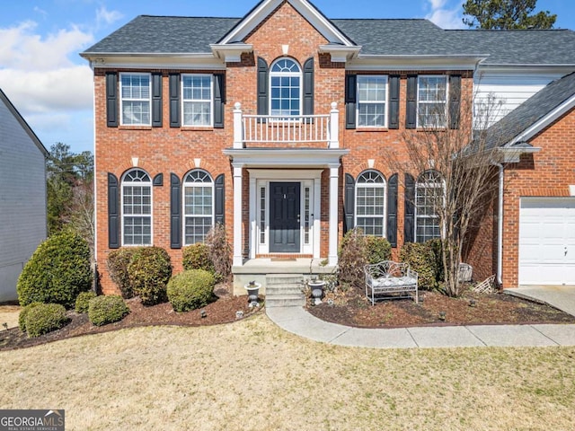 colonial-style house featuring brick siding, a front yard, a balcony, and roof with shingles