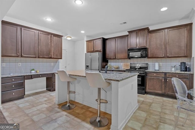 kitchen featuring a breakfast bar area, light stone counters, an island with sink, black appliances, and built in desk