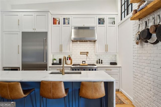 kitchen with a sink, stainless steel built in fridge, light stone counters, and range hood