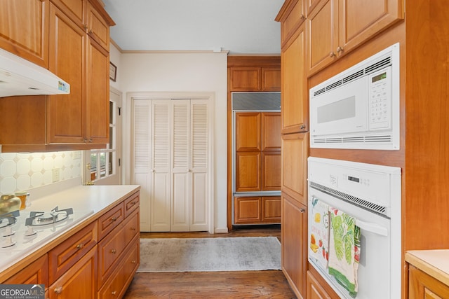 kitchen featuring under cabinet range hood, light countertops, decorative backsplash, brown cabinets, and white appliances