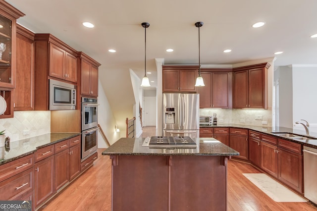 kitchen featuring light wood finished floors, dark stone counters, a sink, hanging light fixtures, and appliances with stainless steel finishes