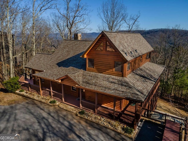 view of front of home with a view of trees, a porch, faux log siding, roof with shingles, and a chimney