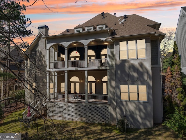 back of property at dusk featuring stucco siding, a balcony, a lawn, and a chimney