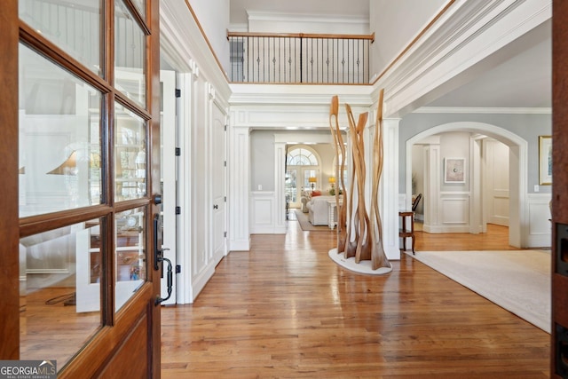 foyer entrance with light wood-style flooring, arched walkways, ornamental molding, a towering ceiling, and a decorative wall