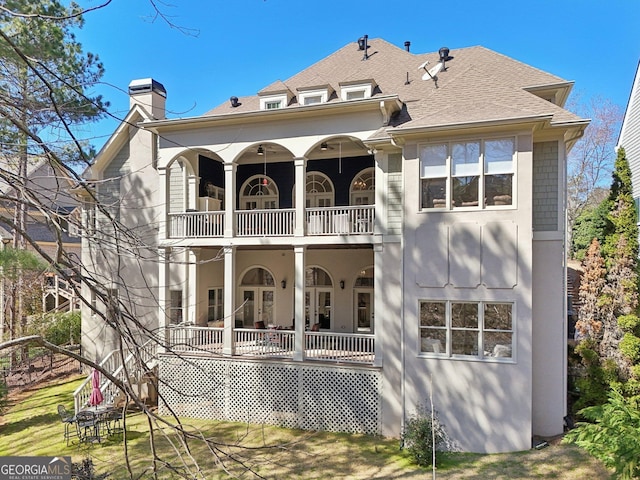 back of house featuring stucco siding, a yard, a shingled roof, a balcony, and a chimney